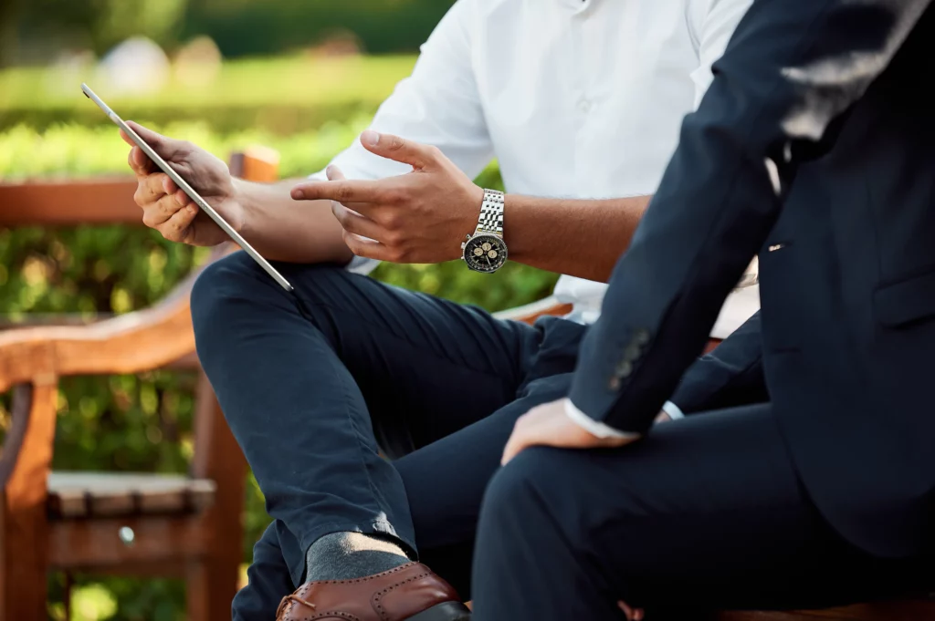 Man sitting on a park bench with a work colleague pointing at the screen of a tablet.